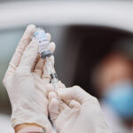 hands with rubber gloves injecting syringe into a glass vial of COVID-19 vaccine with a person blurred in the background