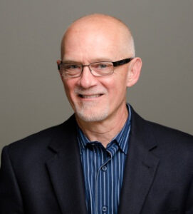 Person with glasses, black jacket and blue striped shirt smiling at camera.