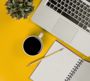 open laptop, coffee cup, notebook, pencil, and plant on top of yellow surface top