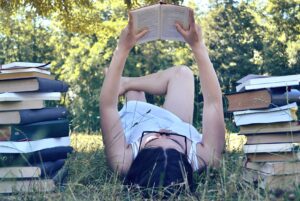 Young female laying down on grass next to a pile of books reading