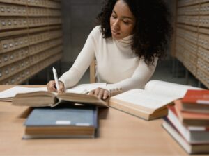 A person with curly hair, wearing a white long-sleeve shirt, sits at a wooden table in a library. They are writing in a notebook with an open book in front of them and several other books stacked nearby. Rows of card catalog drawers are visible in the background.
