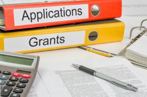 A neatly organized office desk featuring two binders labeled 'Applications' and 'Grants.' The red binder labeled 'Applications' is stacked on top of the yellow binder labeled 'Grants.' A silver pen rests on a document filled with text, while a calculator is positioned to the left. In the background, there are additional documents and an open binder.