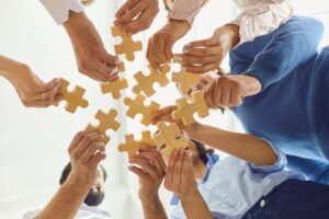 A group of people standing in a circle, each holding a wooden puzzle piece. The image is taken from below, looking up at the individuals as they bring their puzzle pieces together, symbolizing teamwork and collaboration.