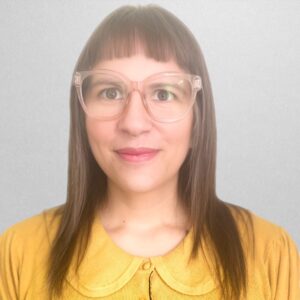 A person with long, straight hair and bangs is wearing large, clear-framed glasses and a mustard yellow top with a rounded collar. The background is plain and light gray.