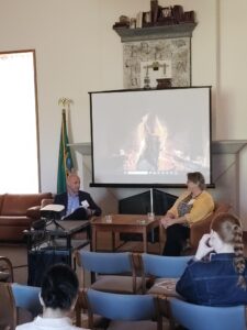 Two people are seated at the front of a room, speaking into a microphone in front of an audience, while others sit nearby, listening attentively. A large screen behind them displays an image of a fireplace.