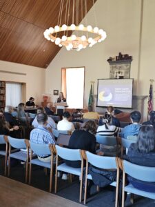 An audience seated in rows facing two speakers at the front of the room, one standing and one sitting behind desks with laptops. A presentation slide on the screen reads "A Global Health Initiative" along with some text and graphics related to health data analysis.