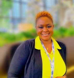 A person with short hair is smiling at the camera. They are wearing a bright yellow shirt, a black jacket, and a lanyard around their neck. The background is blurred, showing greenery and buildings.