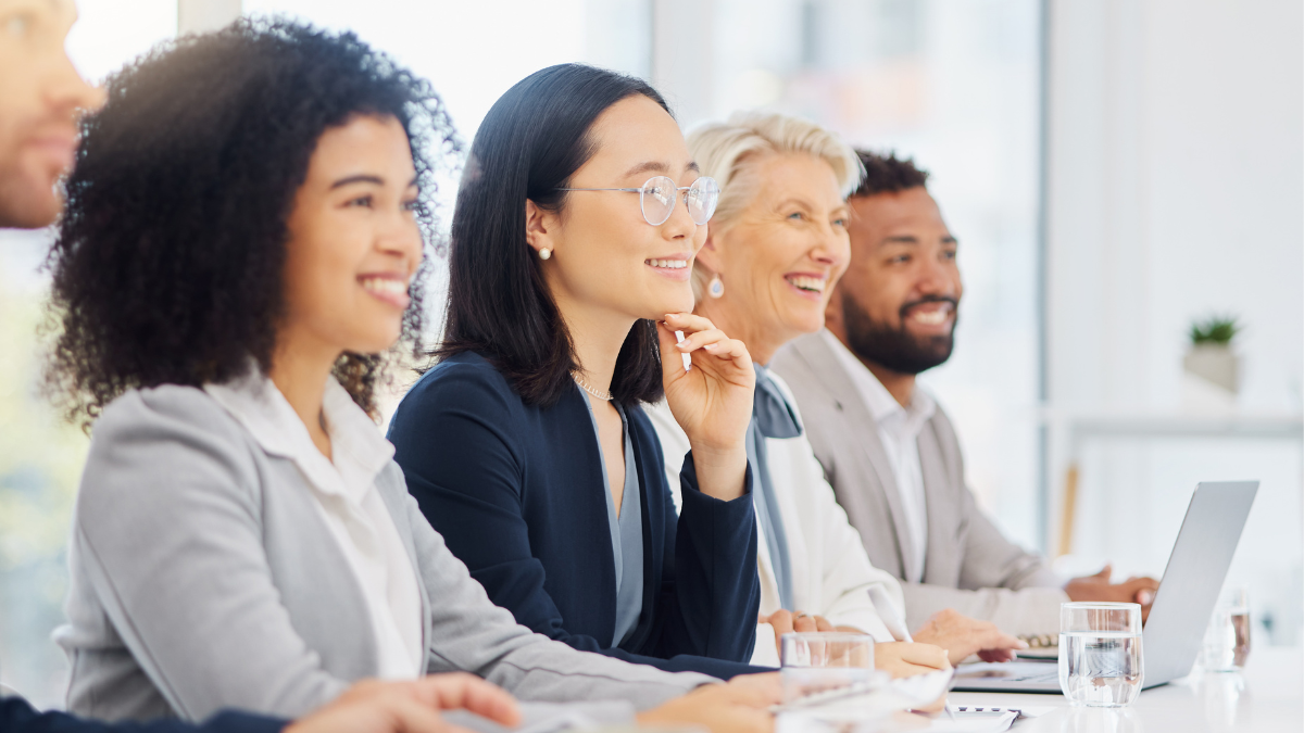A group of people sit side by side at a conference table, looking toward the front of the room while taking notes and working on laptops during a meeting or presentation.