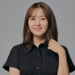 Professional headshot of a woman with shoulder-length brown hair, smiling gently, wearing a short-sleeve black blouse against a light gray background.