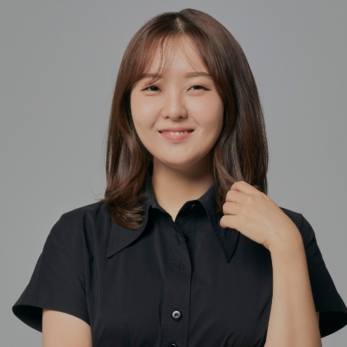 Professional headshot of a woman with shoulder-length brown hair, smiling gently, wearing a short-sleeve black blouse against a light gray background.