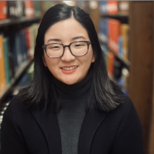 Portrait photo of an individual wearing glasses and a dark blazer, positioned in front of bookshelves filled with books. The background is softly blurred, suggesting a library or study setting.