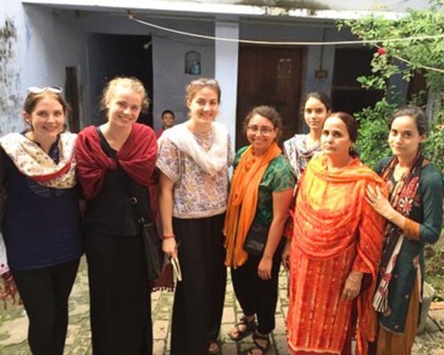 A group of seven people standing together outdoors in a courtyard, wearing a mix of casual and traditional clothing, with building walls, doorways, and greenery in the background.