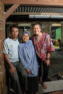 Three people standing together indoors in a covered open‑air structure, with wooden posts and a concrete floor, posing for a photo with additional people and activity visible in the background.