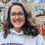 Person wearing a light gray UW sweatshirt standing outdoors beneath blooming cherry trees on the University of Washington campus, with people walking along a path in the background.
