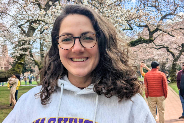 Person wearing a light gray UW sweatshirt standing outdoors beneath blooming cherry trees on the University of Washington campus, with people walking along a path in the background.