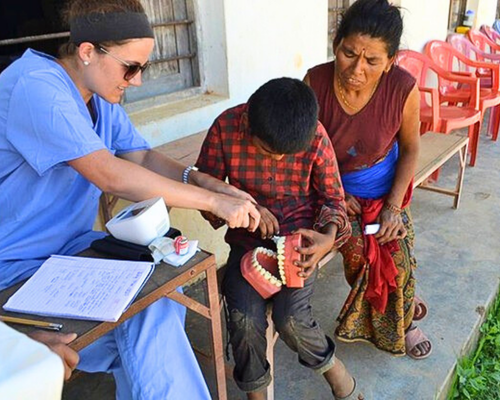 Three people seated outdoors as one person wearing blue scrubs guides another person’s hands while they hold a dental model with removable teeth, with a third person observing beside them; a clipboard and chair are nearby.