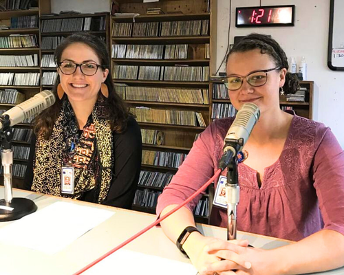 Two people seated at a table in a radio studio, each speaking into a microphone, with shelves of CDs behind them and a digital wall clock displaying the time.