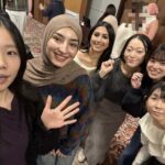 Six smiling people pose for a group selfie in an indoor event space. They are standing close together, looking up at the camera, with patterned carpet and other attendees visible in the background.