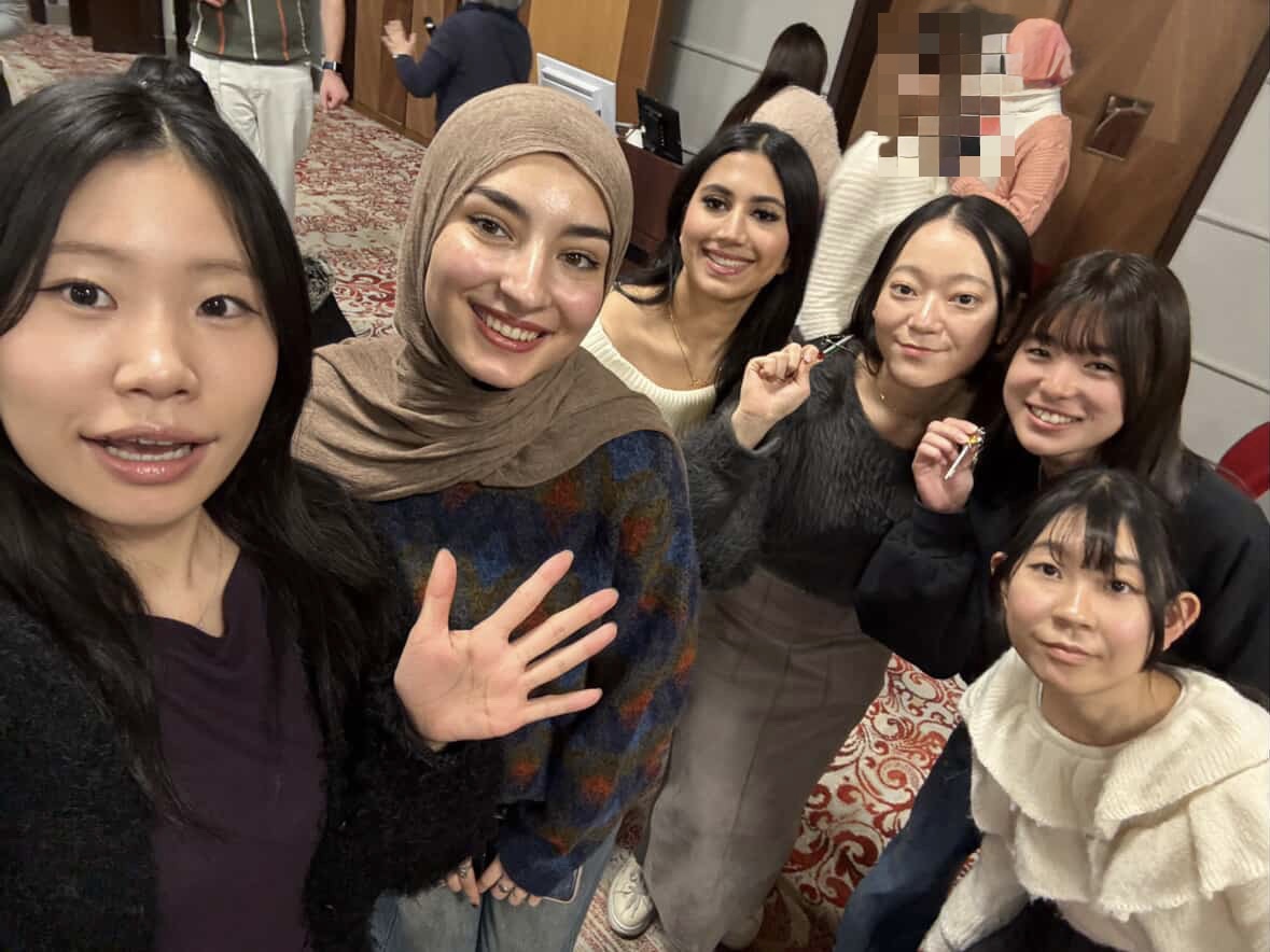 Six smiling people pose for a group selfie in an indoor event space. They are standing close together, looking up at the camera, with patterned carpet and other attendees visible in the background.