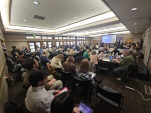 Large audience seated at round tables in a conference room, listening to a speaker presenting at the front near a projected slide.