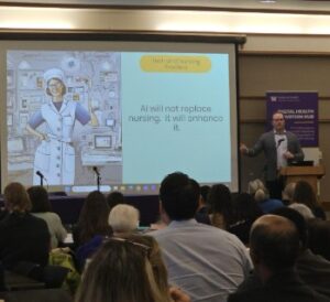 Presenter speaking at a conference with a slide reading “AI will not replace nursing. It will enhance it.