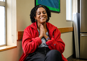 A person seated indoors near a window, wearing a red jacket and dark pants, with hands pressed together at chest level. The person is sitting against a wall with natural light coming in from the left, and a stainless steel refrigerator is visible in the background.