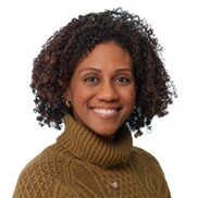 Head-and-shoulders portrait of a person with short, curly hair, wearing a high-collar knit sweater. The background is a plain, light-colored studio backdrop.
