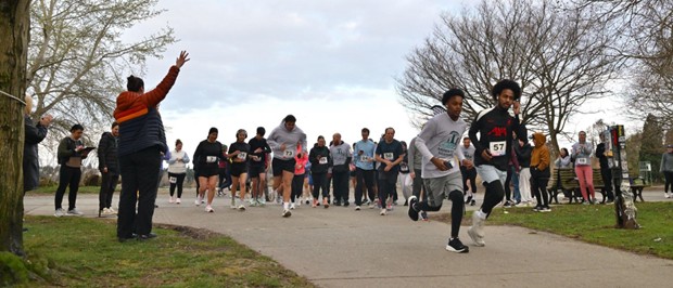 A group of runners begins a race on a paved park path, moving toward the camera. Several runners wear numbered bibs, while a group of people stand along the path and grassy areas nearby. One person at the left raises an arm near the starting area. Leafless trees, grass, and benches are visible under an overcast sky.