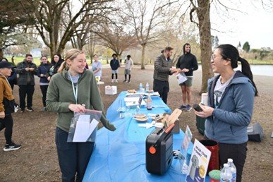 A volunteer table set up outdoors along a park path, covered with a blue tablecloth and supplies. Two people stand facing each other at the table holding papers, while several others gather in the background near leafless trees. Items on the table include water bottles, small containers, and a portable speaker, suggesting an event check-in or support station.