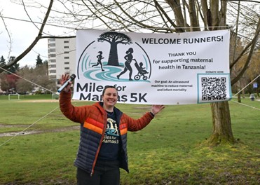  Person standing in a grassy park holding a large banner that reads “Welcome Runners! Thank you for supporting maternal health in Tanzania,” with the “Miles for Mamas 5K” logo, silhouettes of runners and caregivers, and a QR code labeled “Donate here”; trees and apartment buildings appear in the background.