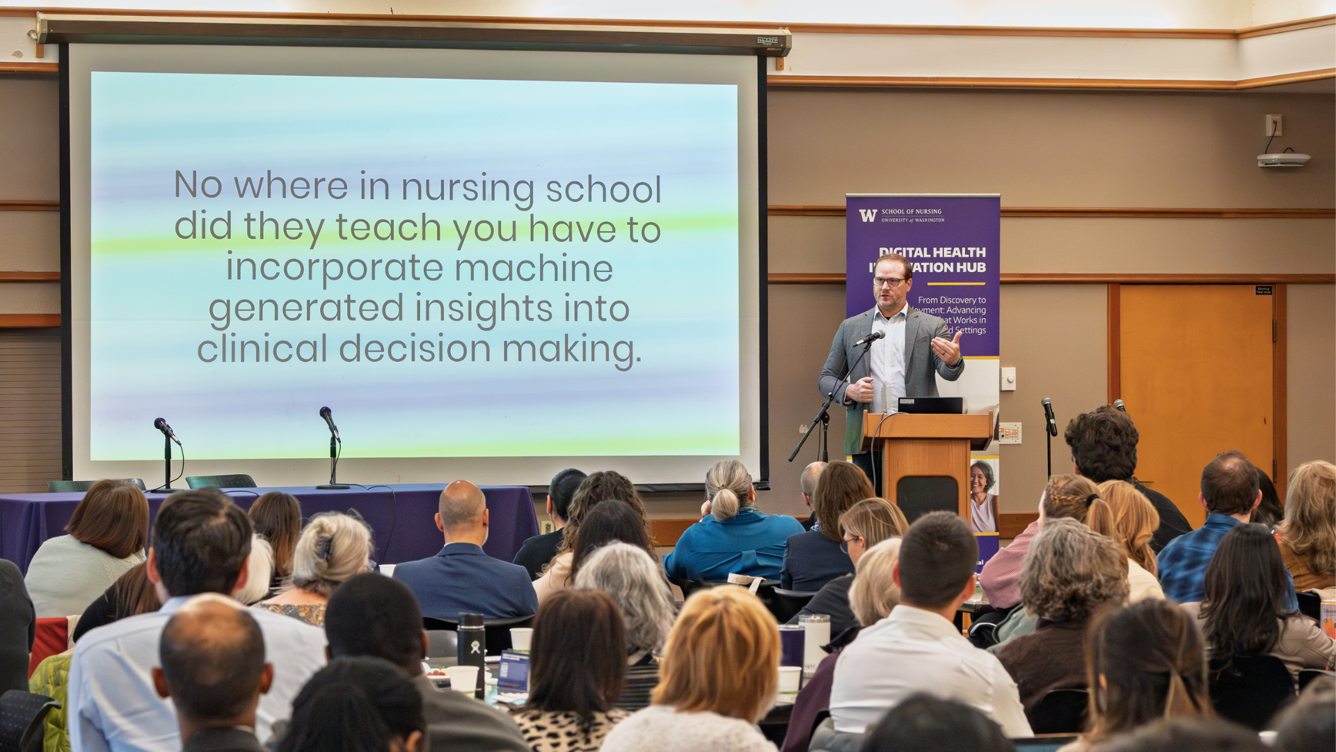 A man at a podium giving a presentation in front of a crowd of people. There are words on the screen that say how generative AI is not taught to be used in clinical decision making.