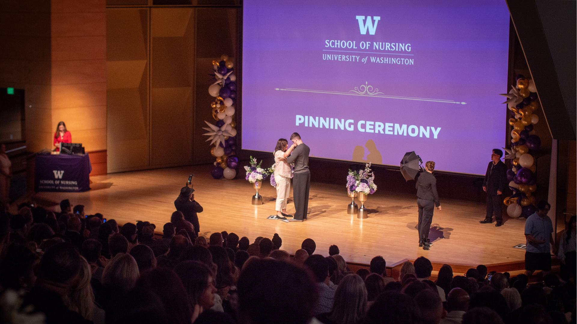 A pinning ceremony taking place on a stage. A woman is putting a pin on a man's suit jacket.