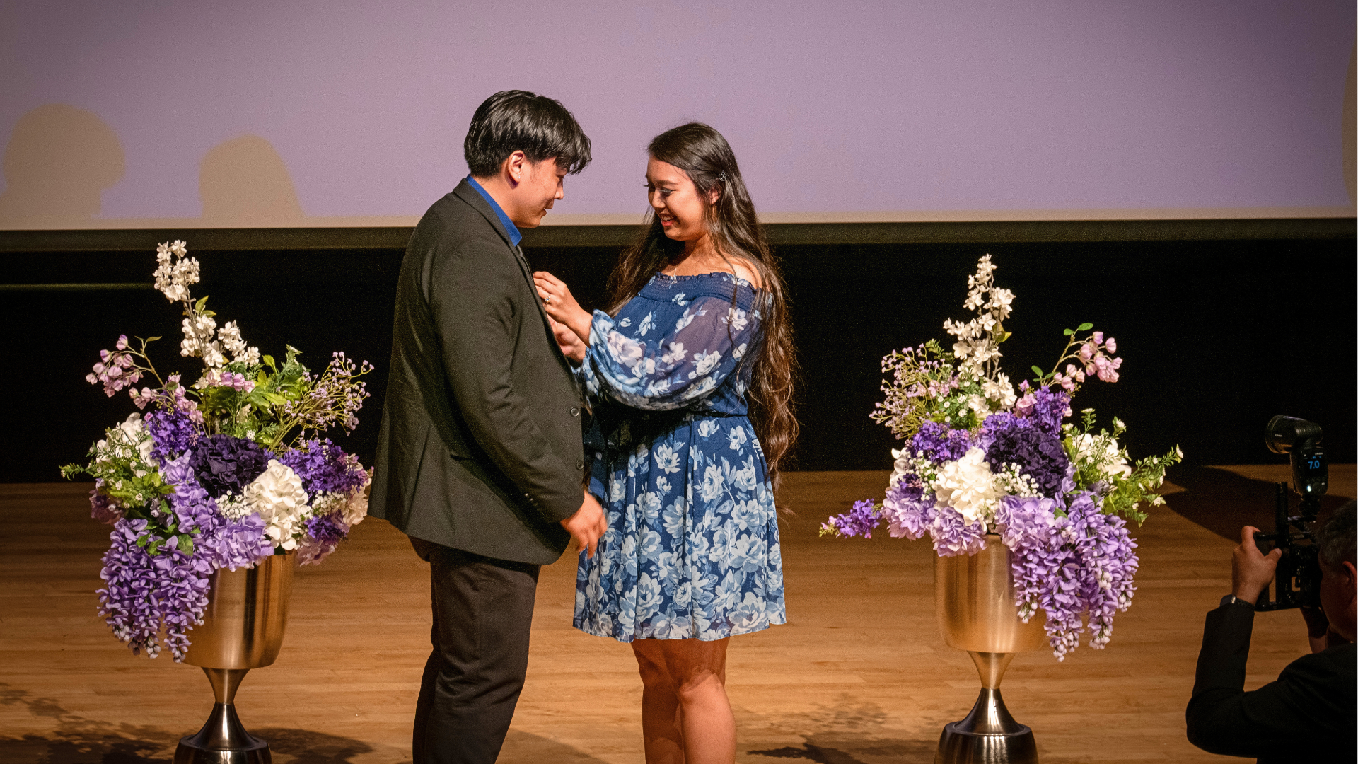 Two people standing on a stage. A man on the left and woman on the right. The woman is putting a pin on the man's suit.