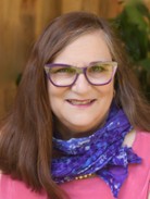 Head-and-shoulders portrait of a person with long brown hair wearing rectangular glasses, a pink top, and a purple patterned scarf, photographed indoors against a softly blurred background.