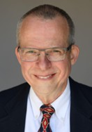Head-and-shoulders portrait of a person with short gray hair wearing glasses, a dark suit jacket, a white shirt, and a red patterned tie, photographed against a plain gray background.