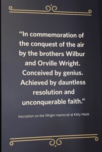Decorative plaque with a quote in white text on a dark background that reads: “In commemoration of the conquest of the air by the brothers Wilbur and Orville Wright. Conceived by genius. Achieved by dauntless resolution and unconquerable faith.” Below, smaller text reads: “Inscription on the Wright memorial at Kitty Hawk.” Ornamental gold accents appear at the top and bottom.