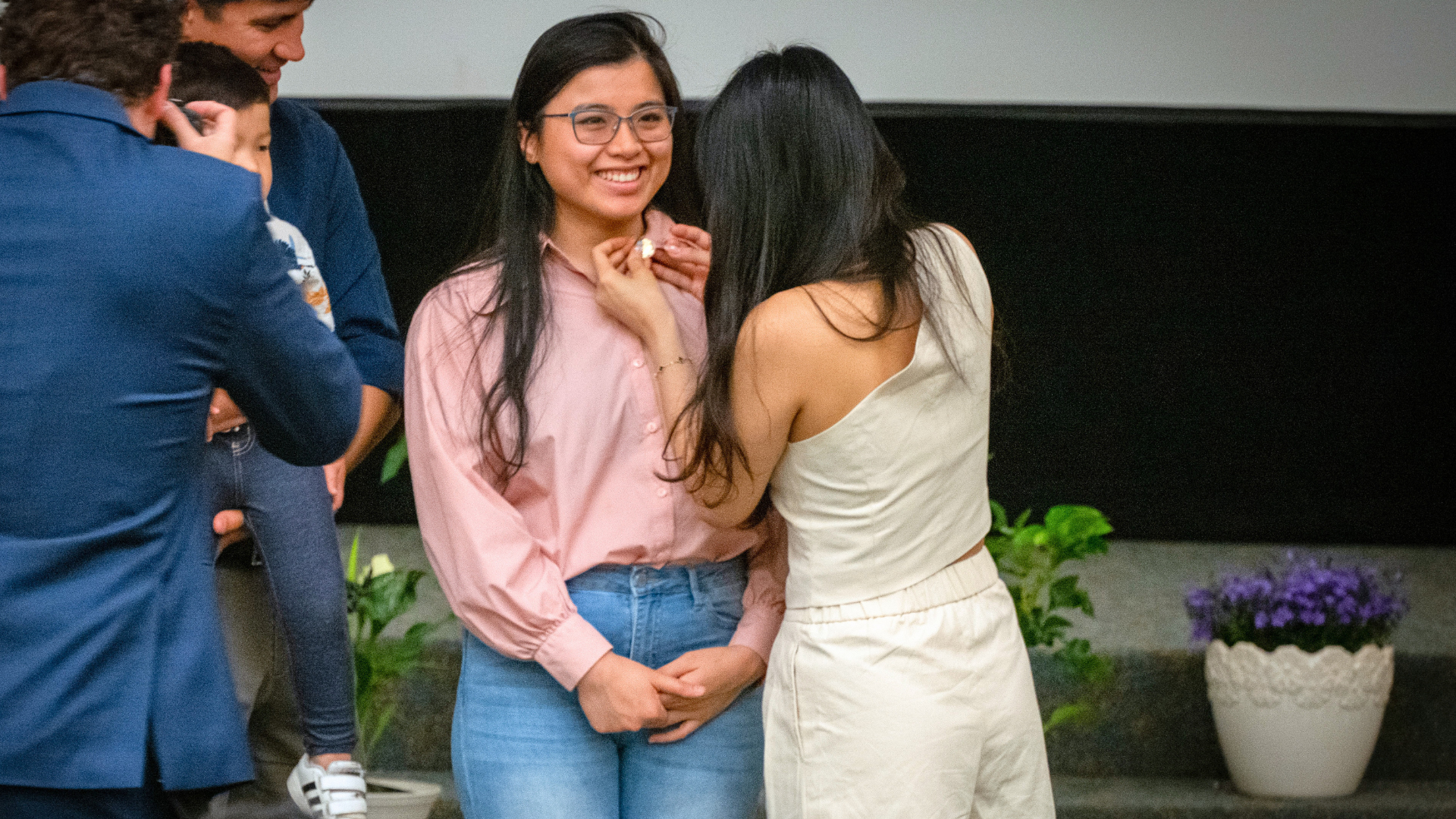 A nursing pinning ceremony taking place on a small stage, where one person is carefully fastening a ceremonial pin onto the shirt of a new nursing graduate wearing a pink button-up shirt and glasses. The graduate stands with hands clasped while others, including someone holding a child, look on from the side. Decorative plants and flowers line the background, emphasizing the formal and celebratory nature of the milestone event.
