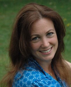 Lydia Professional headshot of a person with long auburn hair wearing a blue patterned blouse, photographed outdoors against a softly blurred green background.