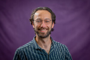 Warren Professional headshot of a person wearing round glasses and a blue striped collared shirt, photographed against a purple studio background.