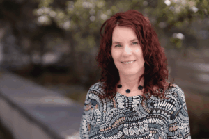 Smiling person with shoulder-length red curly hair wearing a patterned knit top, standing outdoors on a tree-lined path with a soft, blurred background.
