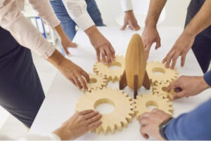 Multiple people collaborate around a table, positioning interlocking wooden gears around a central rocket-shaped model, symbolizing teamwork, coordination, and the collective effort required to advance research and innovation.