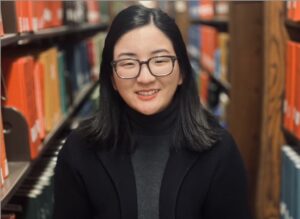 Smiling person with shoulder-length dark hair and black-rimmed glasses, wearing a dark blazer and gray turtleneck, standing in a library aisle between shelves of books.