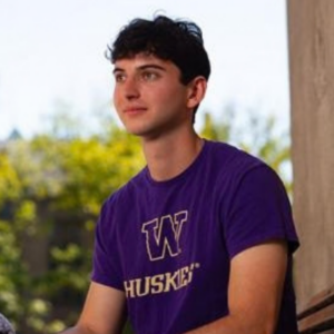 A person in a purple T-shirt sits outdoors on a bench, holding a gray microphone with a foam windscreen while looking to the side; trees and a sunny sky fill the background.