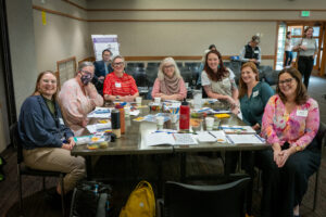 Symposium attendees seated at tables smile and hold up sheets of paper from a group activity.