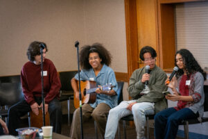 Four students sit together during a musical performance; one plays an acoustic guitar, two hold microphones and one sits on a box drum.